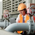 Engineer in safety gear inspecting industrial steel pipelines with a tablet.